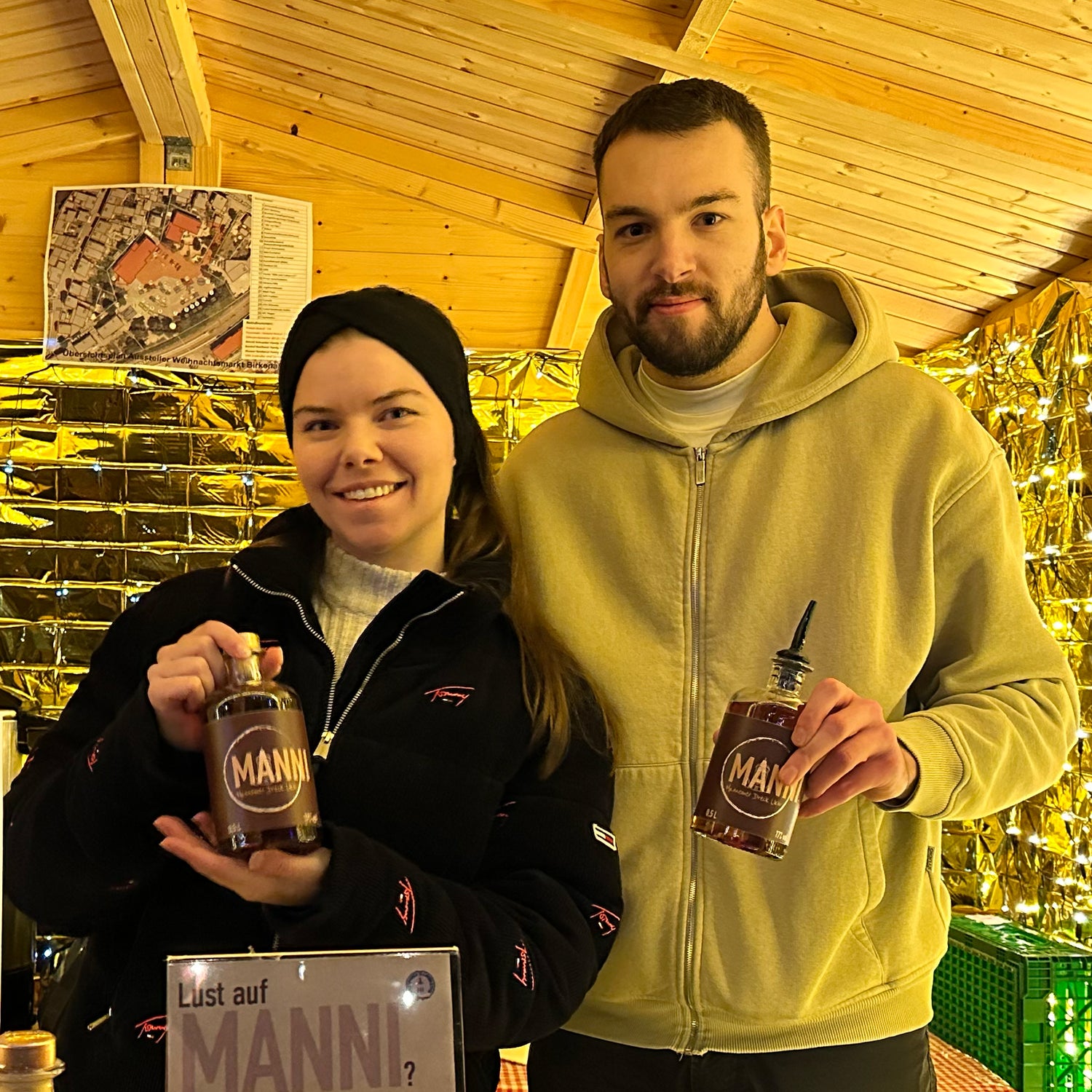 Gründerin und Gründer stehen in einer Weihnachtsmarkthütte und heben Mannemer Dreck Likör Flaschen