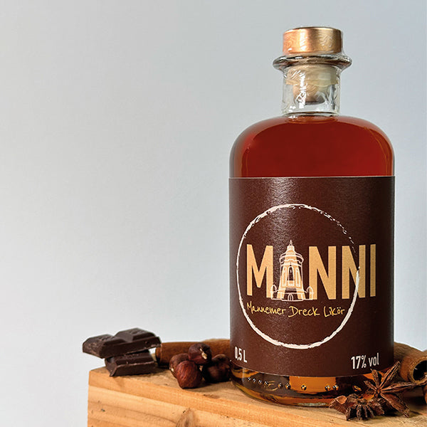 Eine Flasche des Likörs MANNI auf Holzkiste mit Schokolade, Zimt, Nüssen und Anis, grauer Hintergrund