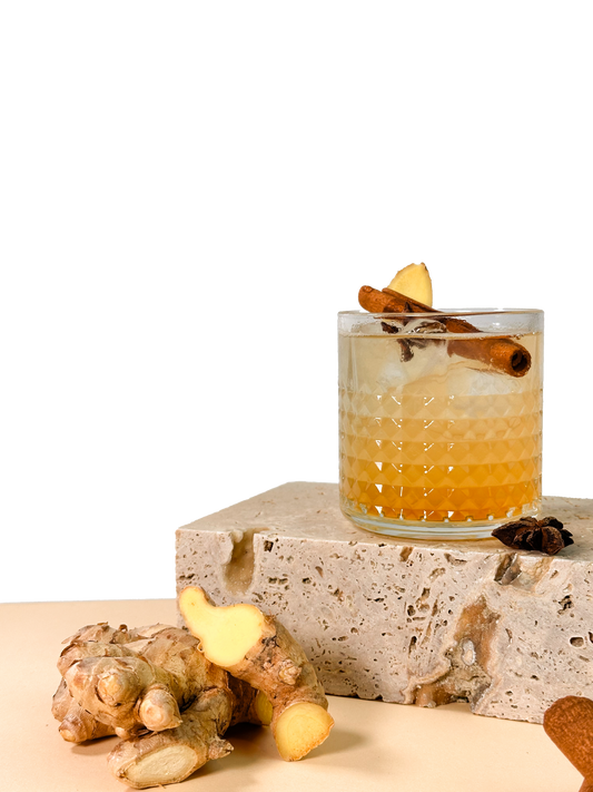 Ein Cocktail mit Ingwerscheibe und Zimtstange in einem Glas auf Stein, daneben eine Ingwerknolle