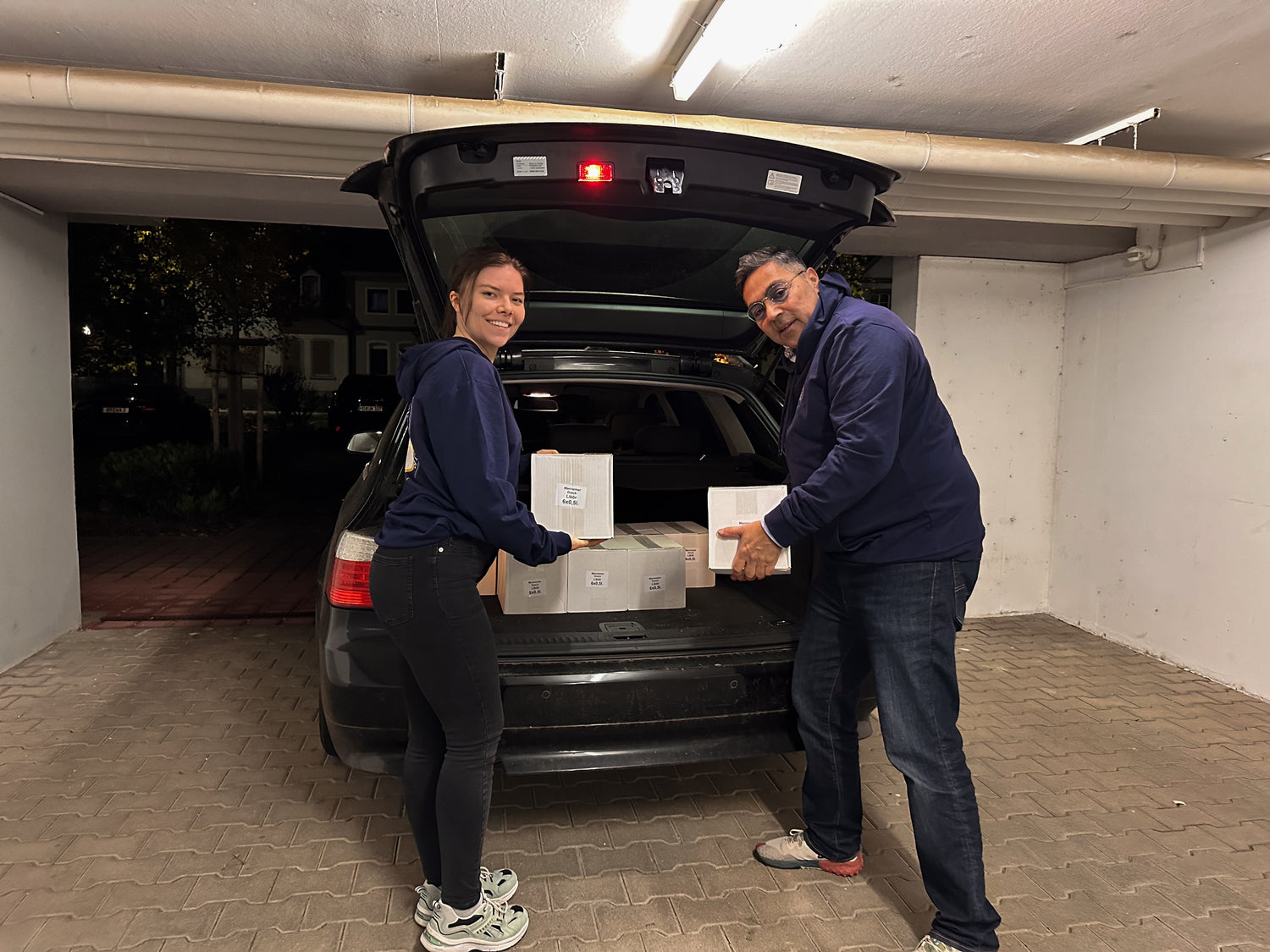 Die Gründerin und der Gründer laden gemeinsam Kisten in ein Auto, abends in der Garage